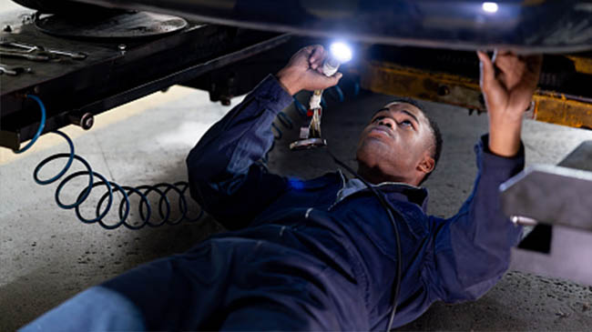 Mechanic in a blue uniform lying under a vehicle, using a flashlight to inspect the undercarriage while working on a car lift.