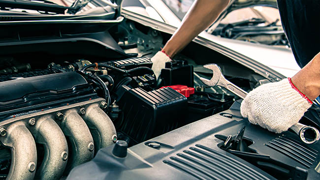 Close-up of hands wearing white work gloves holding a wrench over an open car engine bay, focusing on the engine manifold and battery area.