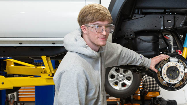 A young mechanic in safety glasses and a grey hoodie working on a brake assembly or wheel hub, with a yellow car lift visible in the background of the auto shop.