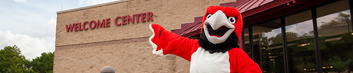 A mascot in a red hawk costume with a large white beak stands with one arm outstretched in a welcoming gesture in front of a Welcome Center building sign
