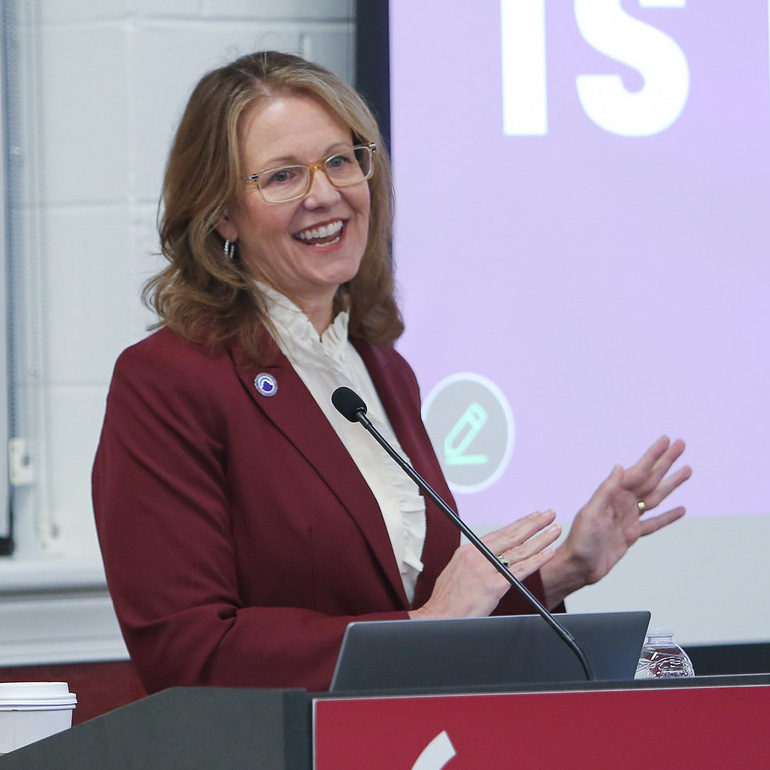 A smiling woman with shoulder-length wavy blonde hair and glasses stands behind a podium, gesturing with both hands while speaking. She is wearing a maroon blazer over a white ruffled blouse and has a small circular pin on her lapel. A microphone is positioned in front of her, and a laptop and water bottle are visible on the podium. In the background, a large screen displays a light purple slide with the word "IS" visible.