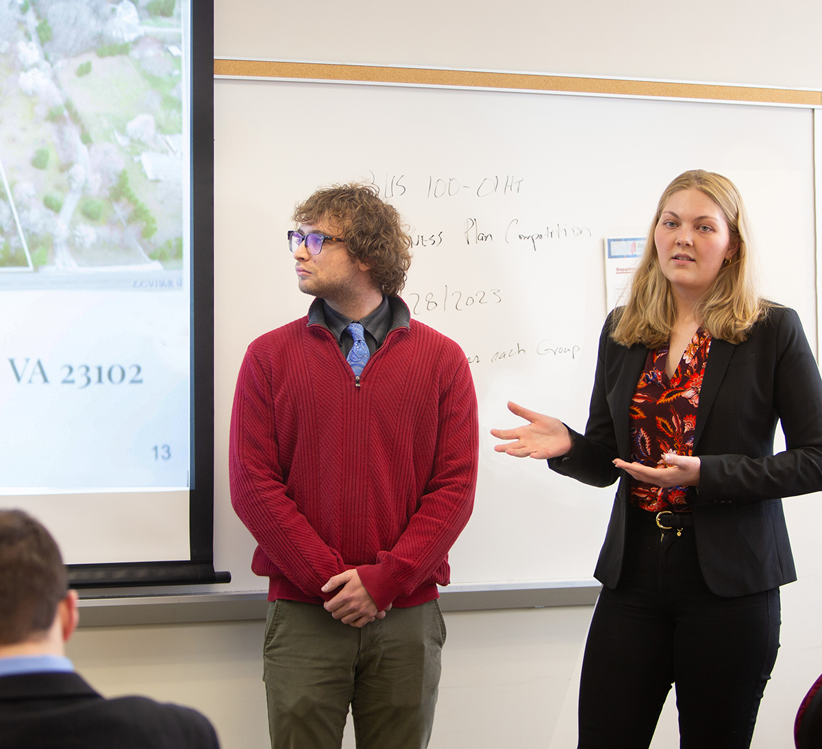 Two students, male in red sweater and female in black pantsuit, stand before the class and pitch their startup business plan