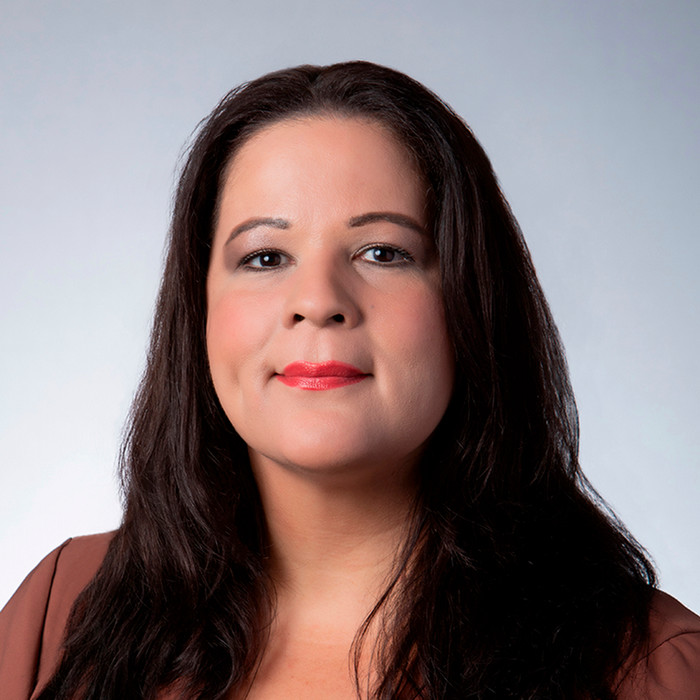 Professional headshot of Carmen Irizarry, Director of Financial Aid at Reynolds Community College. She has long, dark hair parted in the middle and is wearing a brown blazer against a light gray gradient background. She is looking directly at the camera with a neutral, professional expression.