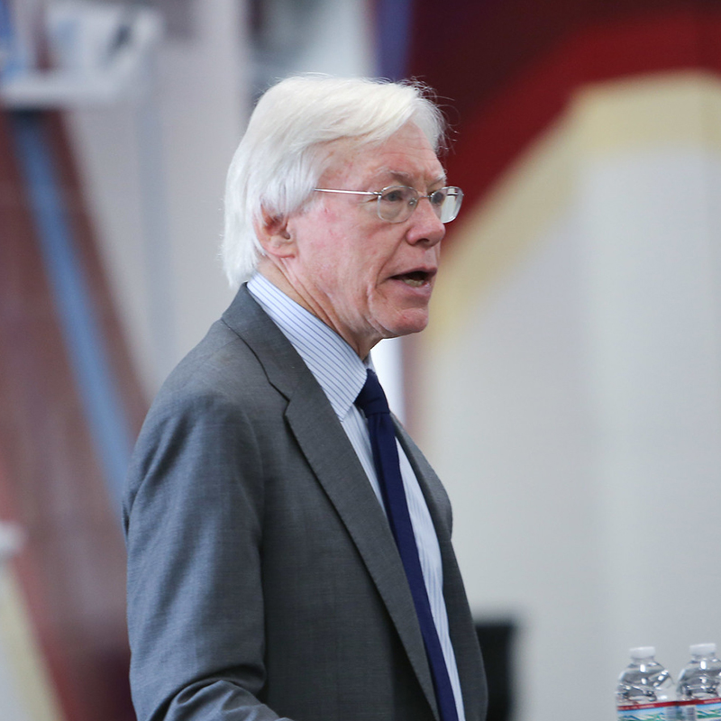 David Baime, an older man with white hair and glasses, speaking at a podium. He is wearing a grey suit jacket, a light-blue pinstriped dress shirt, and a dark navy tie. The background is softly blurred, showing the interior of a bright room at Reynolds Community College.