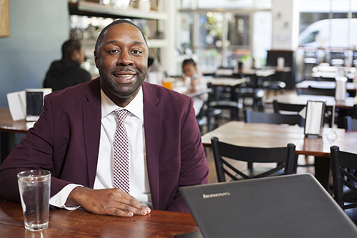 Dominic Brown in burgundy suit and ties smiles at camera as he sits in a restaurant