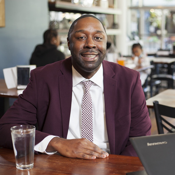 Dominic Brown in burgundy suit and ties smiles at camera as he sits in a restaurant
