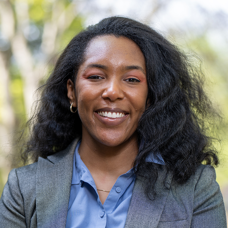 A medium headshot of Ekene Tharpe, a Black woman with a warm, confident smile, outdoors. She has long, voluminous dark hair and is wearing a blue button-down shirt layered under a tailored gray blazer. Her makeup includes a subtle orange-toned eyeshadow, and she is wearing small silver hoop earrings. The background is a soft-focus, natural greenery with dappled sunlight, creating a professional yet approachable aesthetic.