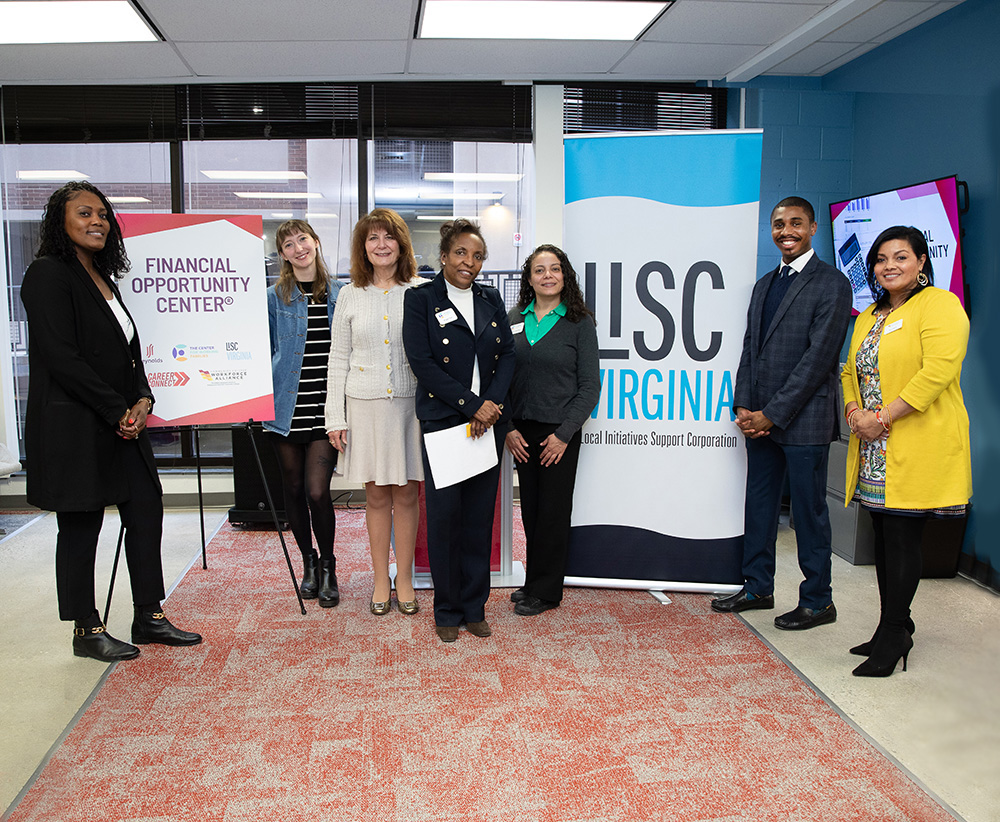 A group of seven diverse professionals stands together in an office setting for a promotional photo. They are posed between two large signs: one on the left for the Financial Opportunity Center® and a large vertical banner on the right for LISC Virginia (Local Initiatives Support Corporation). The individuals are dressed in business and business-casual attire, including blazers, sweaters, and a bright yellow cardigan. They are standing on a patterned red and grey carpet. In the background, large windows show an outdoor area, and a television screen displays financial graphics. The atmosphere is professional and collaborative.