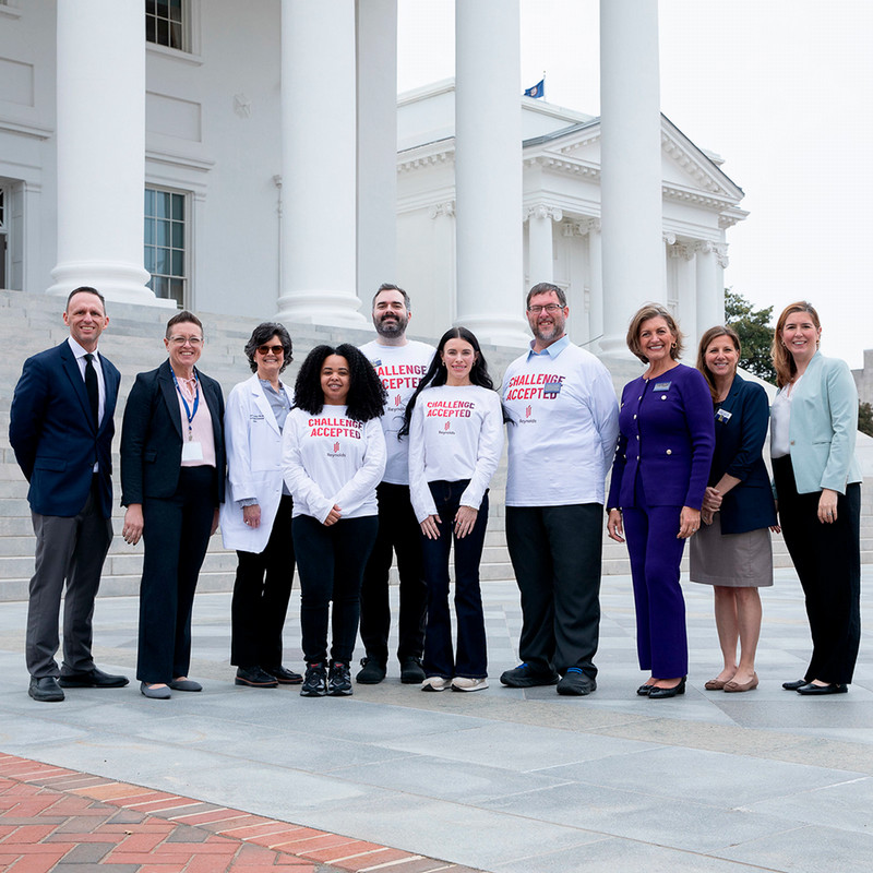 An outdoor group portrait of ten people standing in front of the white columns of a capital building. Four individuals in the center wear white long-sleeved shirts with the red text "CHALLENGE ACCEPTED" and a Reynolds logo. They are flanked by others in professional attire, including a woman in a purple suit and a woman in a white lab coat. The group is smiling and posed on a stone plaza.