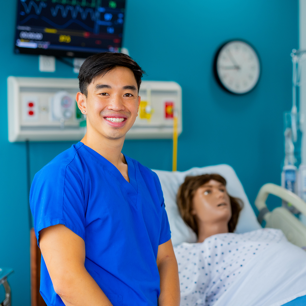 A smiling young man in bright blue medical scrubs stands in a healthcare simulation lab. In the background, a medical manikin lies in a hospital bed wearing a patient gown. The room features teal walls, a wall-mounted heart rate monitor displaying vital signs, a large analog clock, and medical gas outlets.