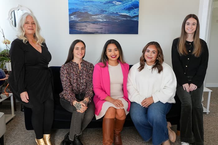 Five women smiling together indoors in front of a white wall with a large blue abstract painting. On the far left, a woman with long blonde hair stands wearing a black dress and metallic boots. Three women sit on a black couch in the center: one in a patterned blouse, Jem Iwatsubo in the middle wearing a bright pink blazer over a neutral dress with knee-high brown boots, and another in a white sweater and blue jeans. On the far right, a woman with long brown hair stands wearing a black cardigan and dark trousers.