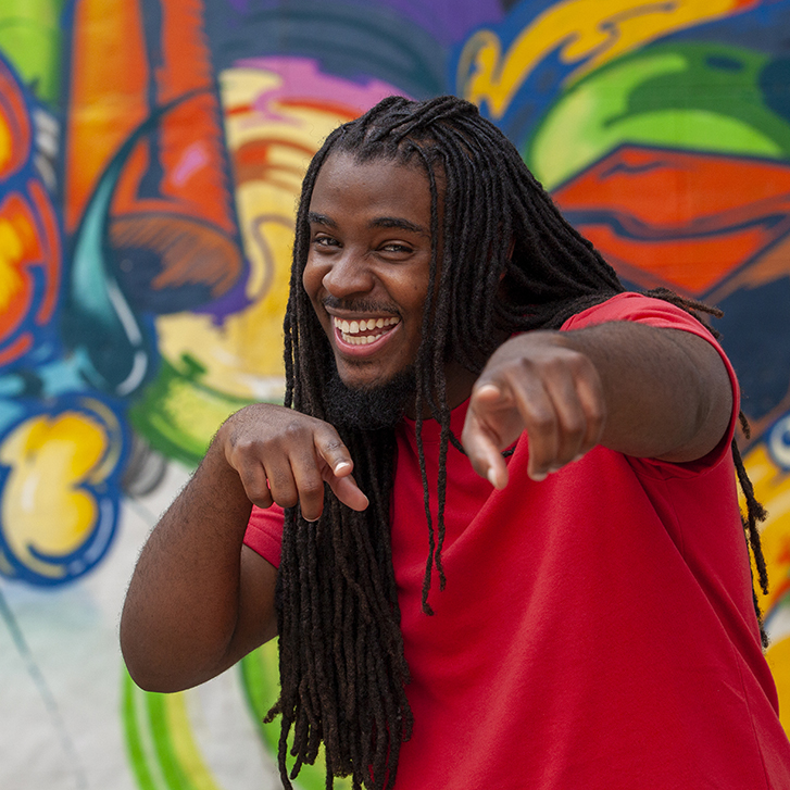Young black man in braids and red shirt playfully points at camera against a colorful graffiti background.