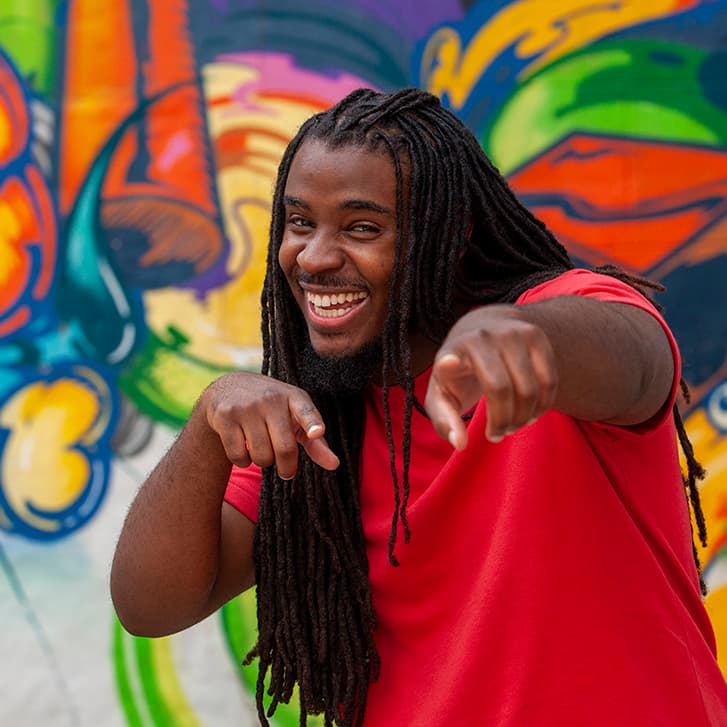 Young black man in braids and red shirt playfully points at camera against a colorful graffiti background.