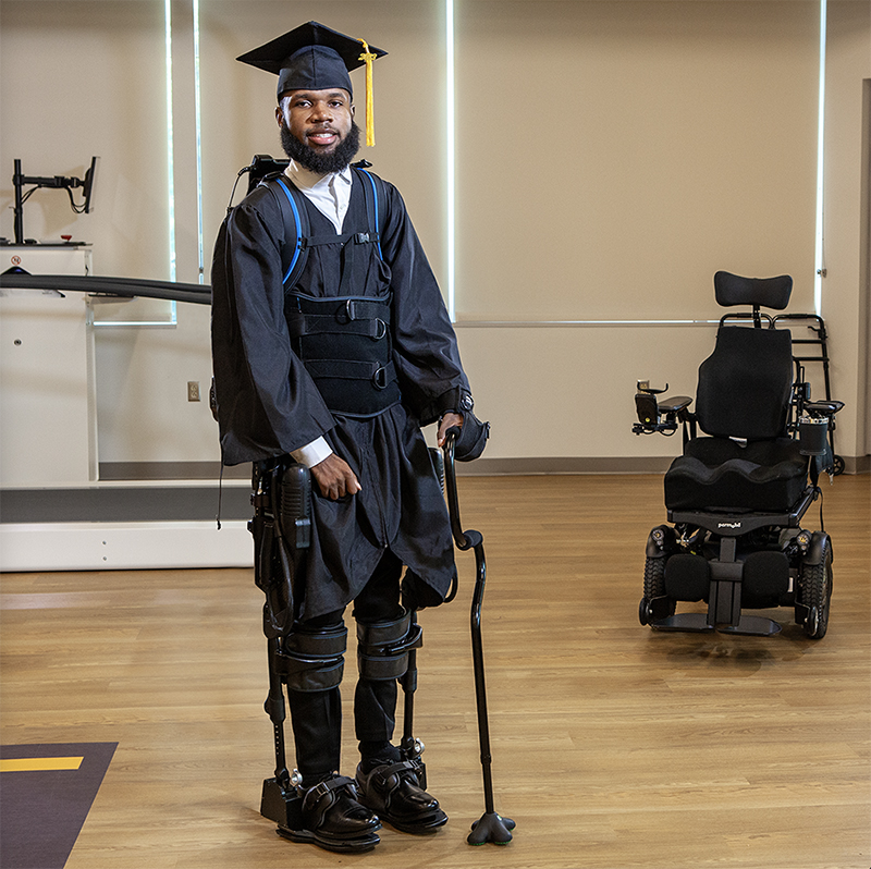 Khalil Watson wearing his graduation cap and gown, stands with the help of exoskeleton