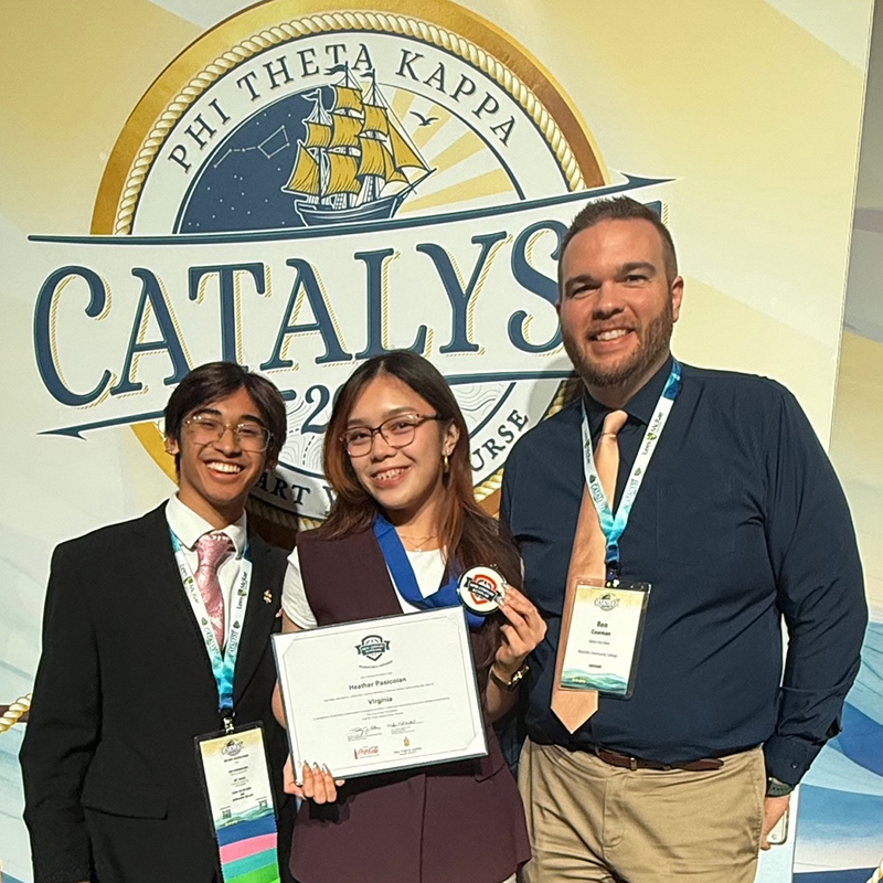 Three people pose for a photo in front of a Phi Theta Kappa "Catalyst" backdrop with a nautical theme. To the left, a young man in a black suit, pink tie, and glasses smiles brightly. In the center, a young woman in a maroon vest and glasses holds an award certificate and a medal. To the right, a man in a navy blue shirt and tan tie stands with his arm near the woman. All three wear conference lanyards. The backdrop features a large circular logo with a sailing ship and the words "Phi Theta Kappa Catalyst."