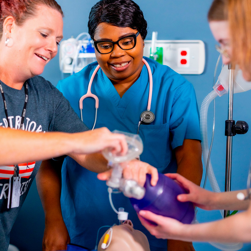 Three healthcare students or professionals in a clinical setting practice using a manual resuscitator (BVM) on a medical mannequin. A woman in a blue scrub top and pink stethoscope smiles while observing two others coordinate the placement of the oxygen mask and bag.