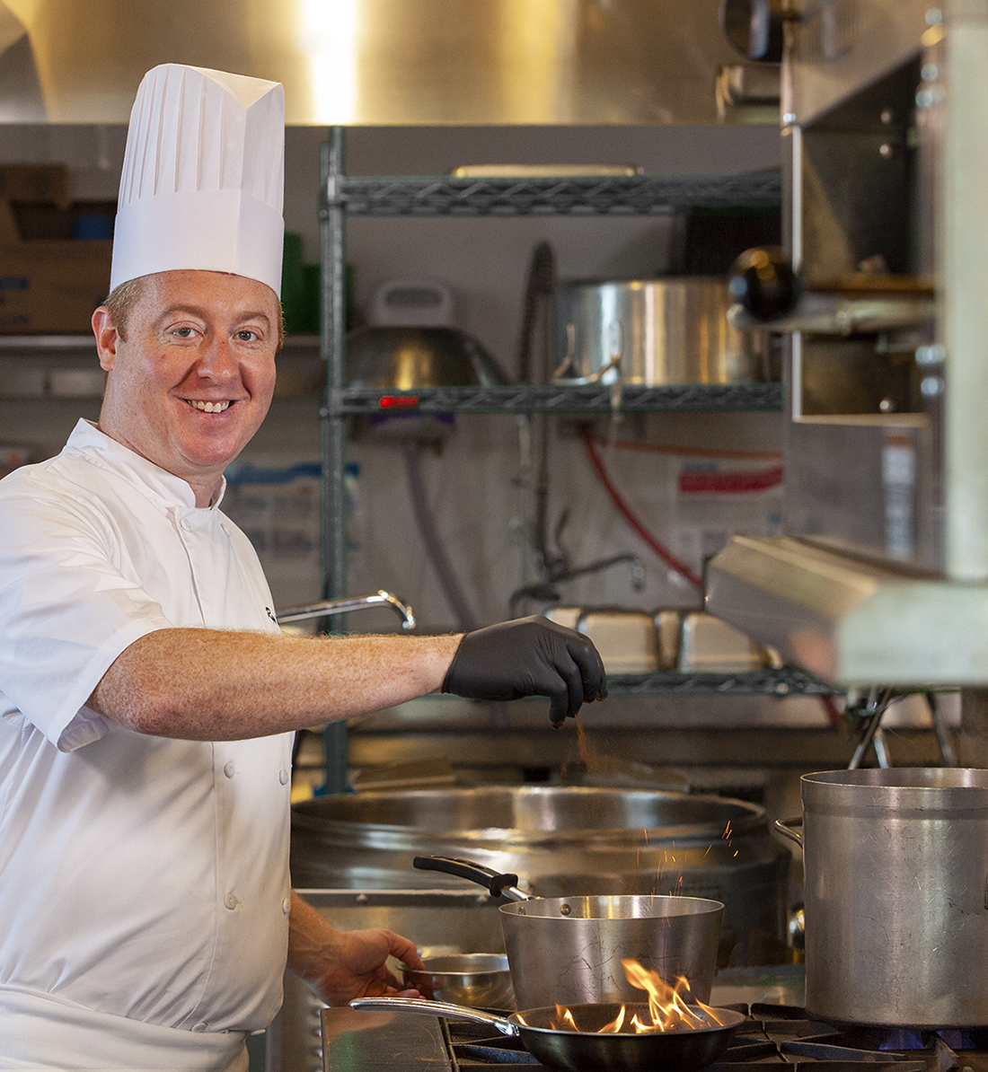 Chef Ryan Manning at the stove in an industrial kitchen in white apron and tall white chef's hat, sprinkling ingredients into a pot