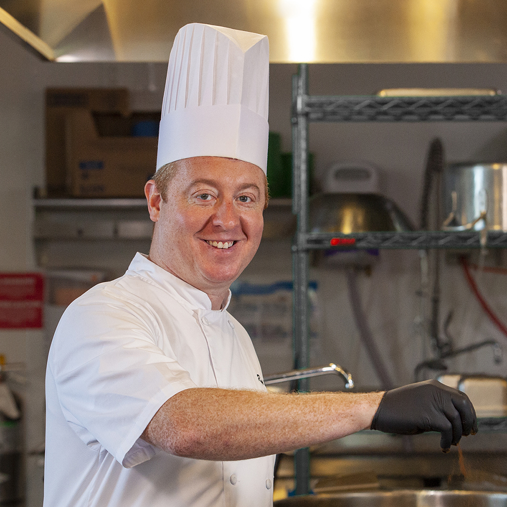 Chef Ryan Manning at a stove in an industrial kitchen wearing tall white chef's hat and apron