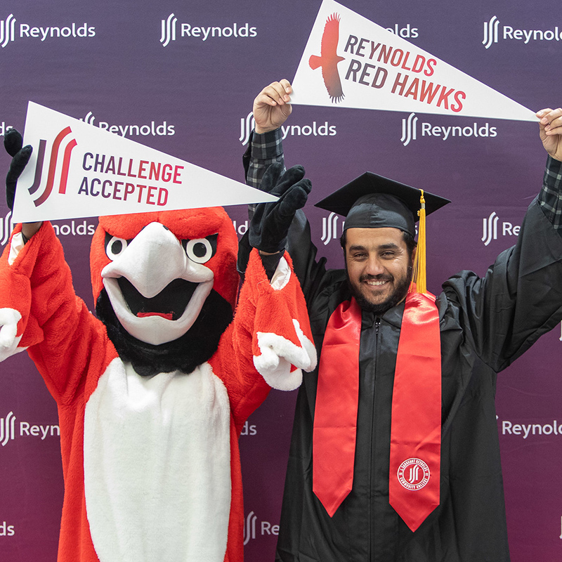 A celebratory photo of Sayed Ahmad Shah Sekandary and Sarge the Red Hawk, the Reynolds mascot, standing against a purple "Reynolds" branded backdrop. Sayed, on the right, wears a black graduation cap and gown with a red stole and smiles broadly while holding up a white pennant that reads "Reynolds Red Hawks." Sarge, a large red and white bird mascot, stands to the left with its wings raised, holding a similar pennant that says "Challenge Accepted." Both are posed energetically to celebrate the graduation achievement.