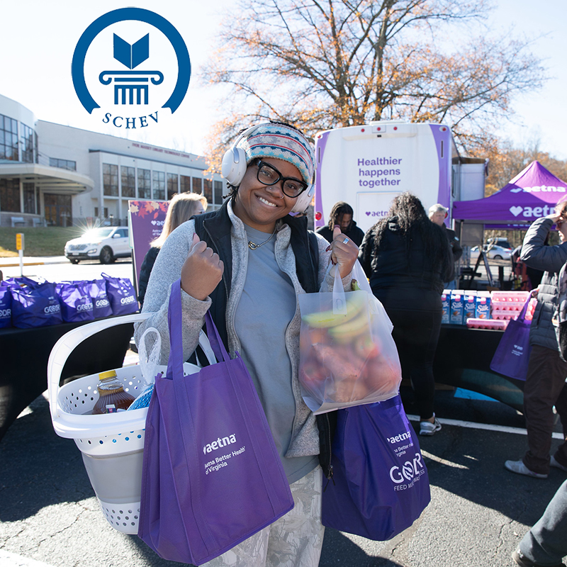A smiling Reynolds Community College student stands outdoors holding several bags of fresh groceries and a laundry basket of supplies. She is wearing headphones and a winter hat, giving a thumbs-up. In the background, an "Aetna Better Health of Virginia" mobile unit and purple tents are set up for a food distribution event. The SCHEV (State Council of Higher Education for Virginia) logo is overlaid in the top left corner.