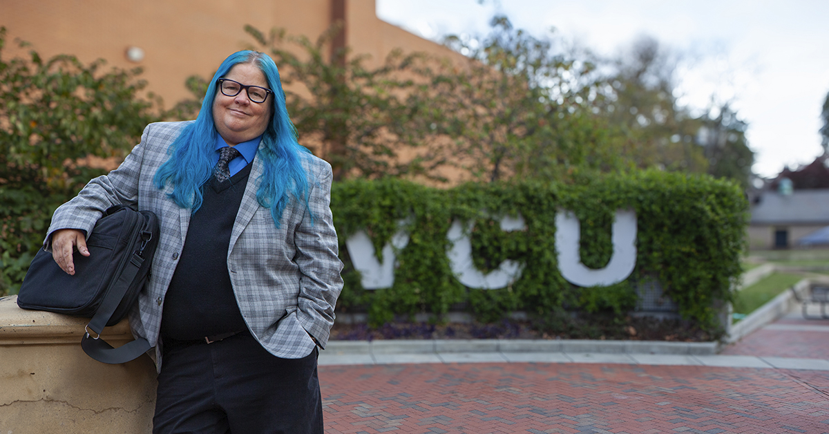 Steph Cull with long blue hair and blue checked jacket smiles at the camera in front of green VCU exterior