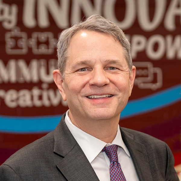 A headshot of Dr. Steve Robinson, a middle-aged man with short, graying hair and a warm smile. He is wearing a dark gray suit jacket, a crisp white dress shirt, and a red patterned tie. The background is a blurred red wall featuring the words "INNOVATE," "EMPOWER," and "COMMUNITY."