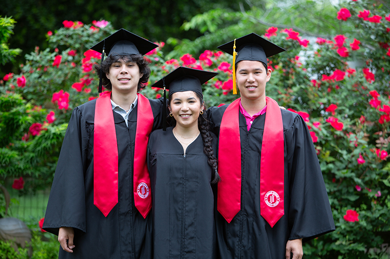 Inocensio siblings pose in their graduation regalia - black caps, graduation gowns and red sashes, in a rose garden