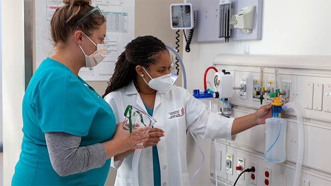 A female healthcare professional in scrubs sits at a desk, using a laptop while surrounded by medical diagrams.