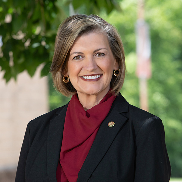 Professional headshot of a woman in a dark gray business suit and cream-colored blouse. She has shoulder-length wavy gray-blonde hair, is wearing small hoop earrings and a pearl pendant necklace, and is smiling warmly at the camera. The background shows red banners and trees on the Reynolds Parham Road campus.